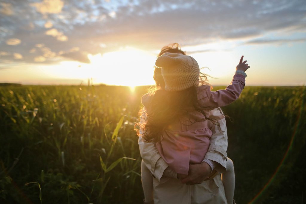 Mother with daughter on back walking through a field of green vegetation at sunset.