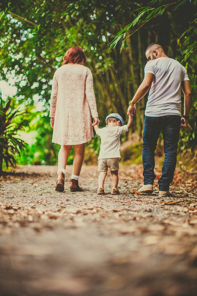 Parents walking with toddler down a path with leaves and green trees.