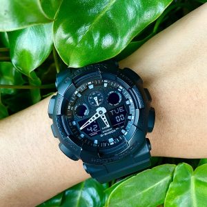 Black digital watch with a black band on a mans wrist surrounded by green foliage.