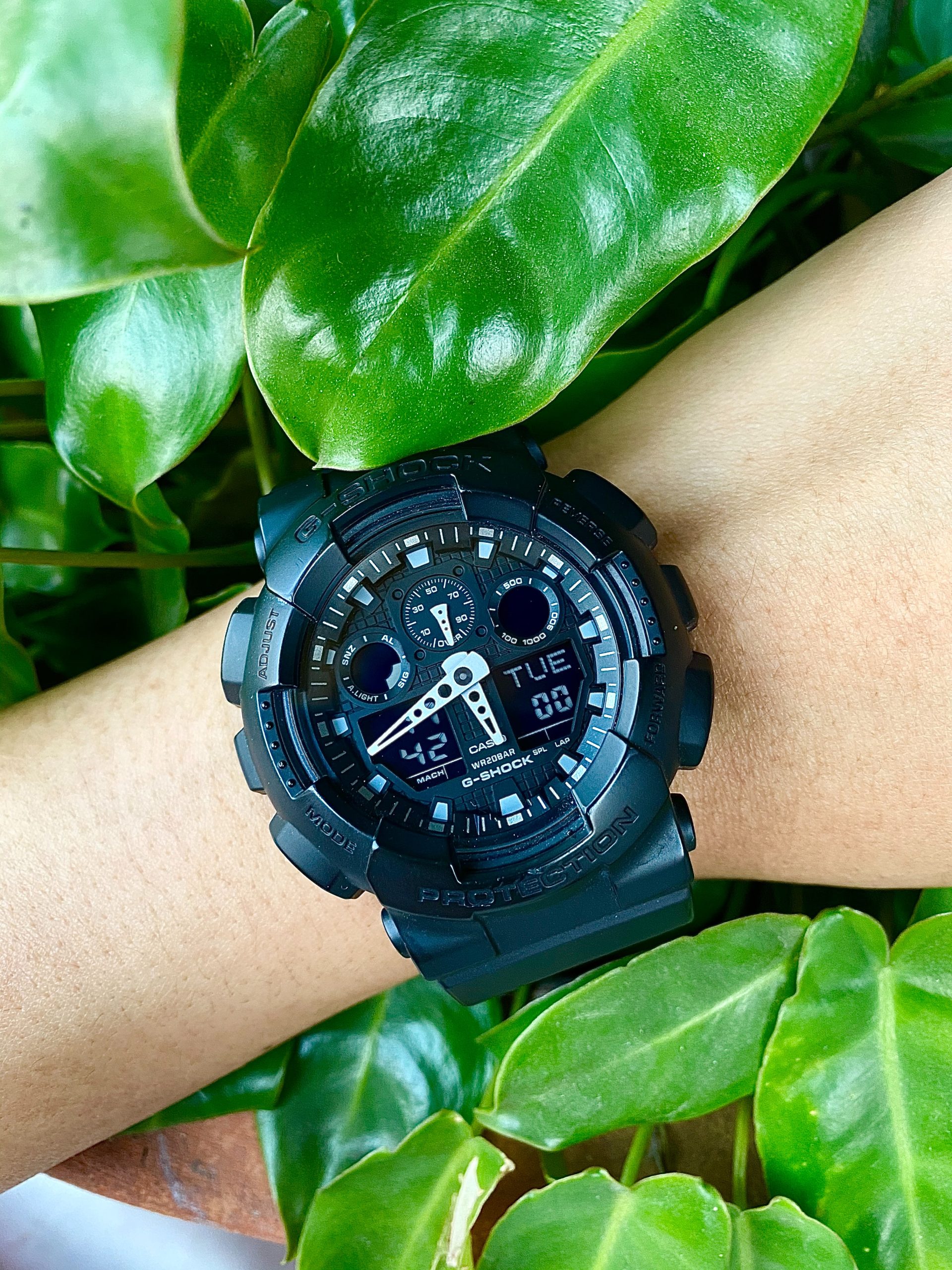 Black digital watch with a black band on a mans wrist surrounded by green foliage.
