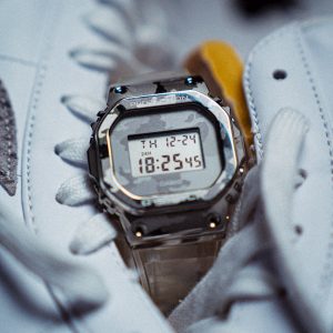 A digital watch by Casio designed with a camouflage band and case sits nestled between to bright white hight top sneakers