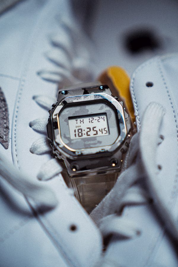 A digital watch by Casio designed with a camouflage band and case sits nestled between to bright white hight top sneakers