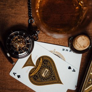 Pocket watch on a table next to a glass of golden liquor and playing cards.