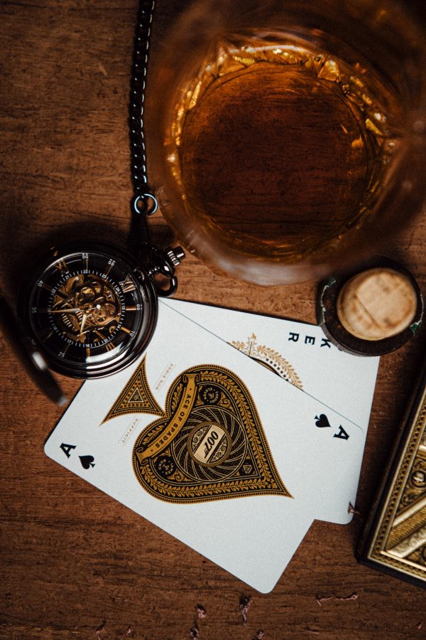 Pocket watch on a table next to a glass of golden liquor and playing cards.