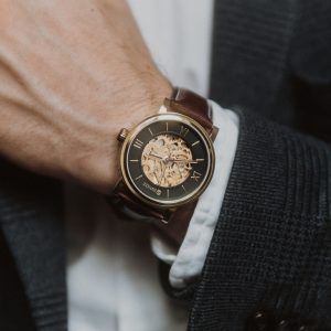 An automatic analog watch with a skeleton key view of the innerworkings, blue and gold face, gold dial, and a brown leather strap sitting on a mans wrist who is wearing a black suit and a white shirt.