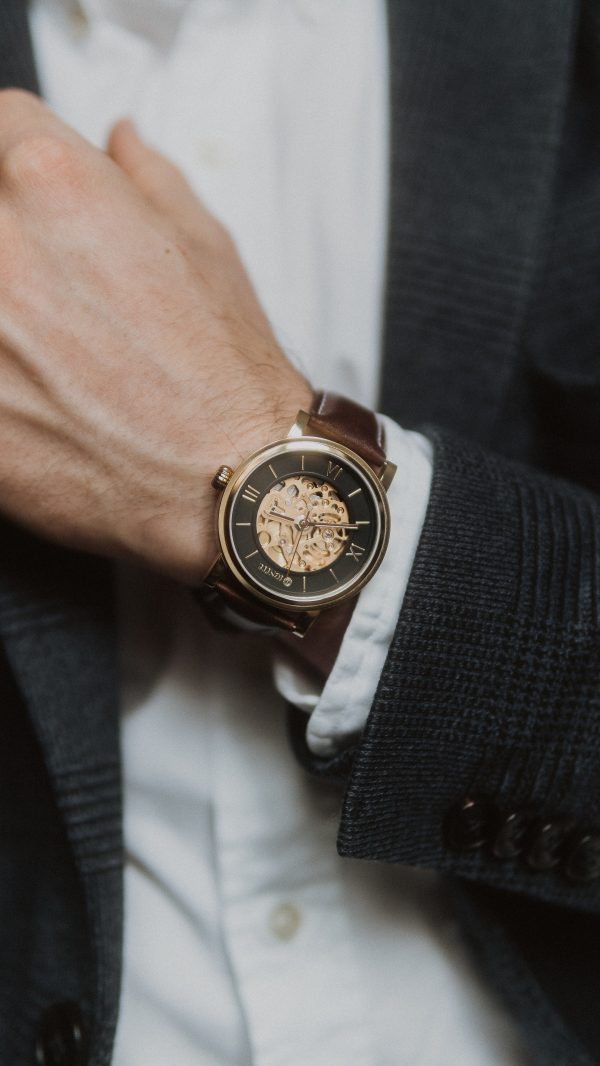 An automatic analog watch with a skeleton key view of the innerworkings, blue and gold face, gold dial, and a brown leather strap sitting on a mans wrist who is wearing a black suit and a white shirt.