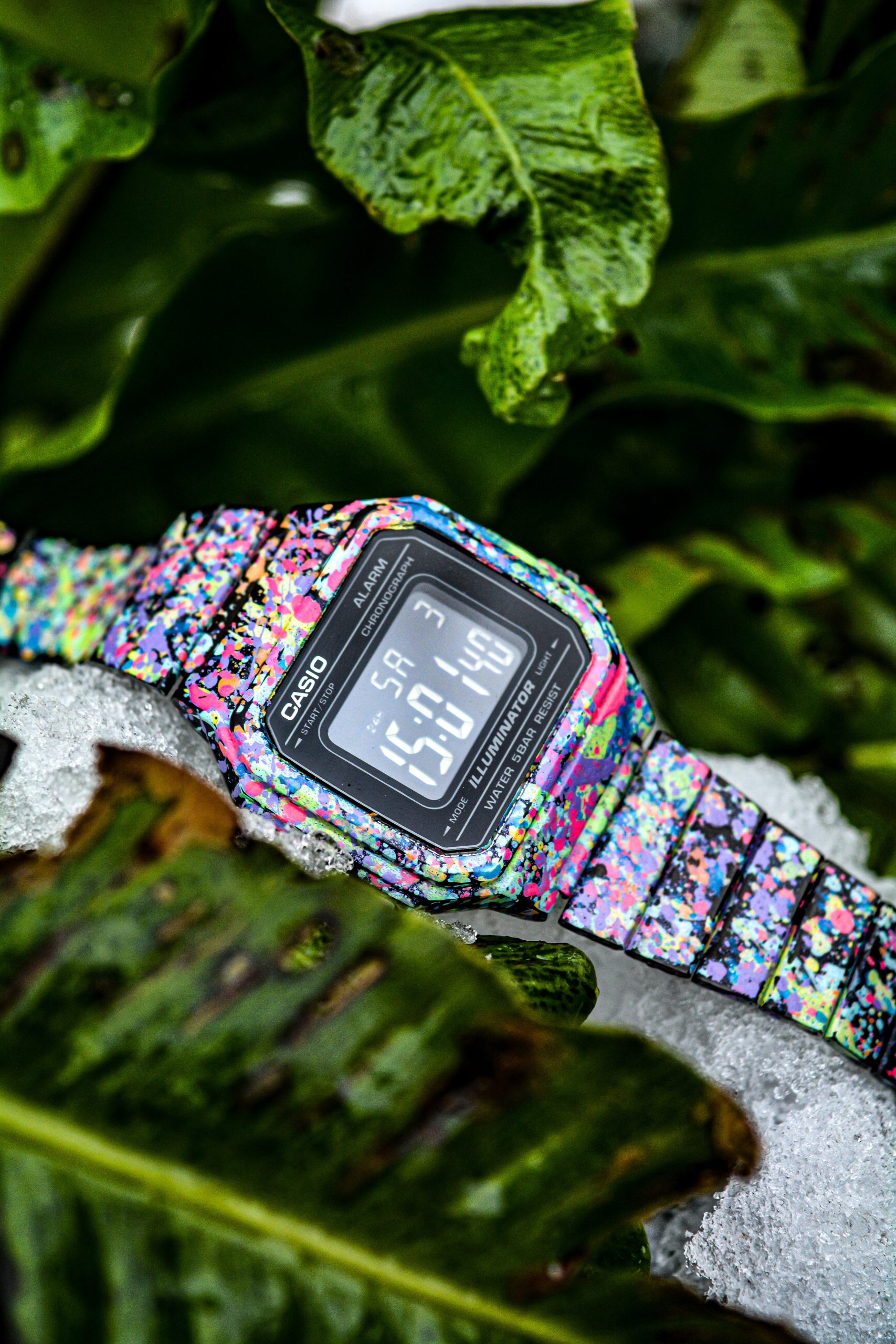 Black digital watch with a black band on a mans wrist surrounded by green foliage.